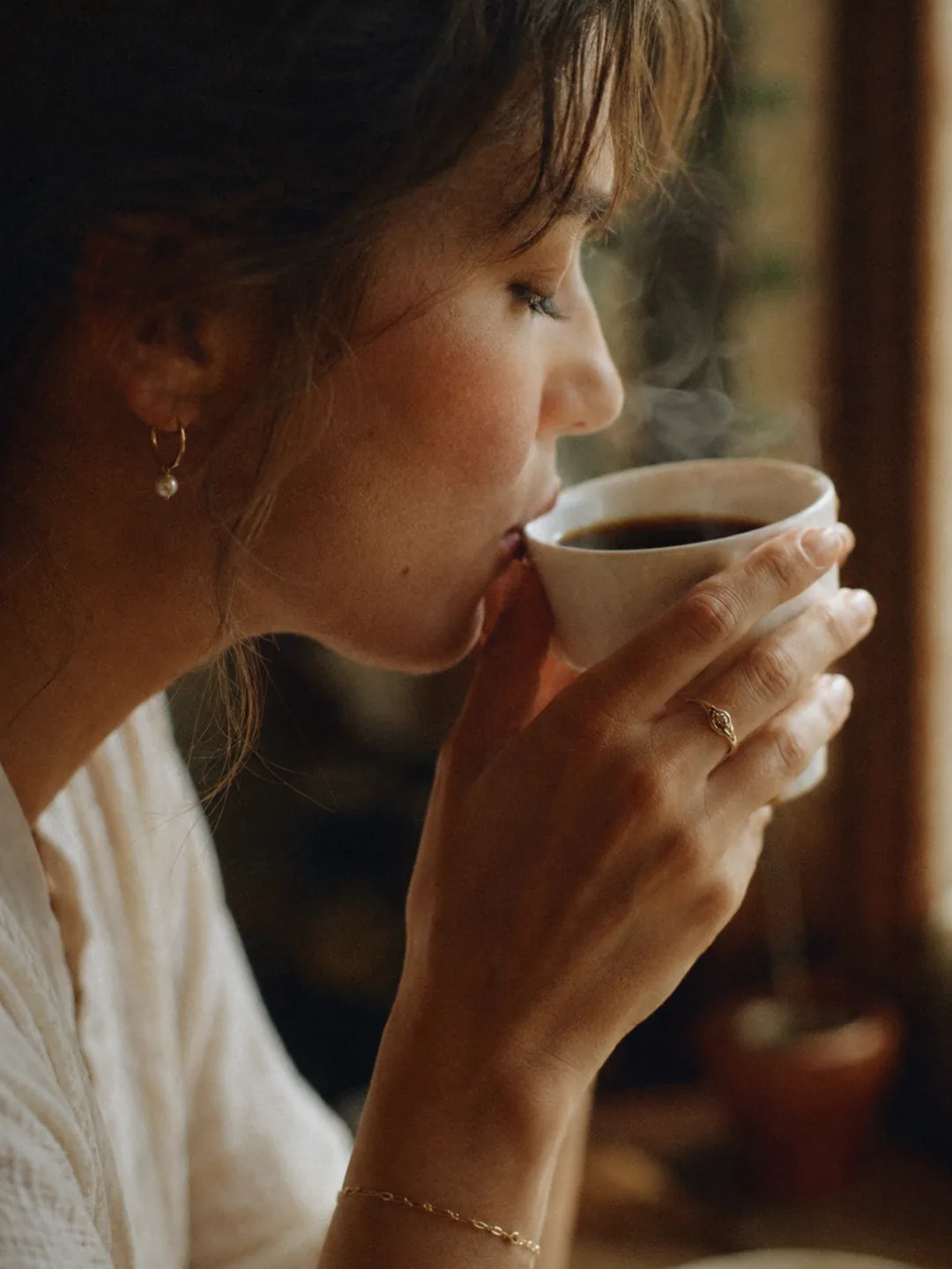 A woman sipping a steaming cup of black coffee by a window
